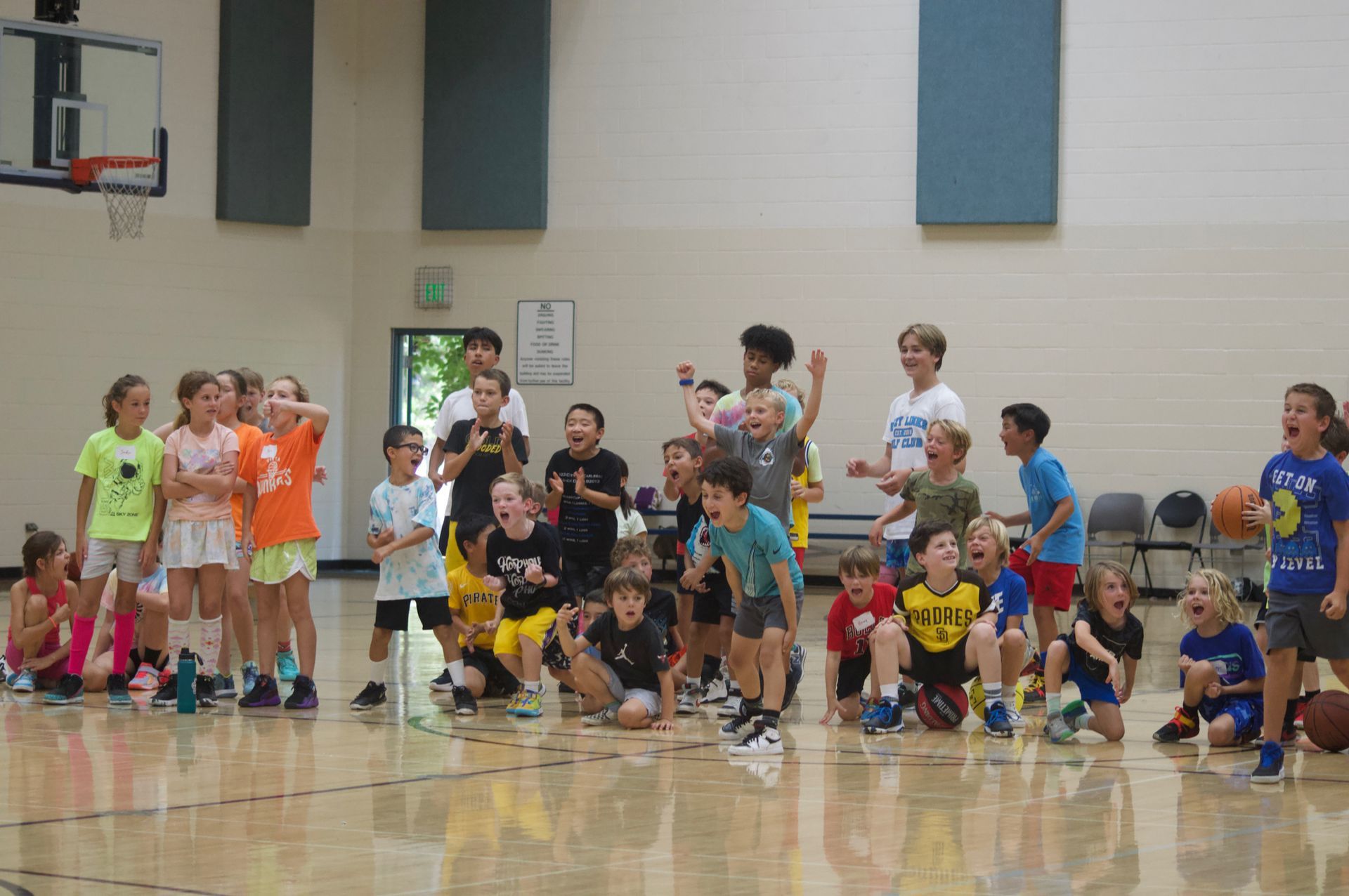 Children crouch in a school gym before a basketball game, with coaches and players lined up behind them.