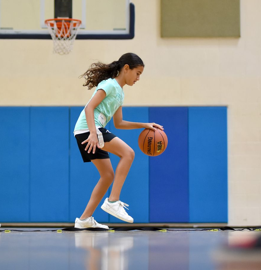 Children dribbling basketballs in a gym during practice, with a coach observing.