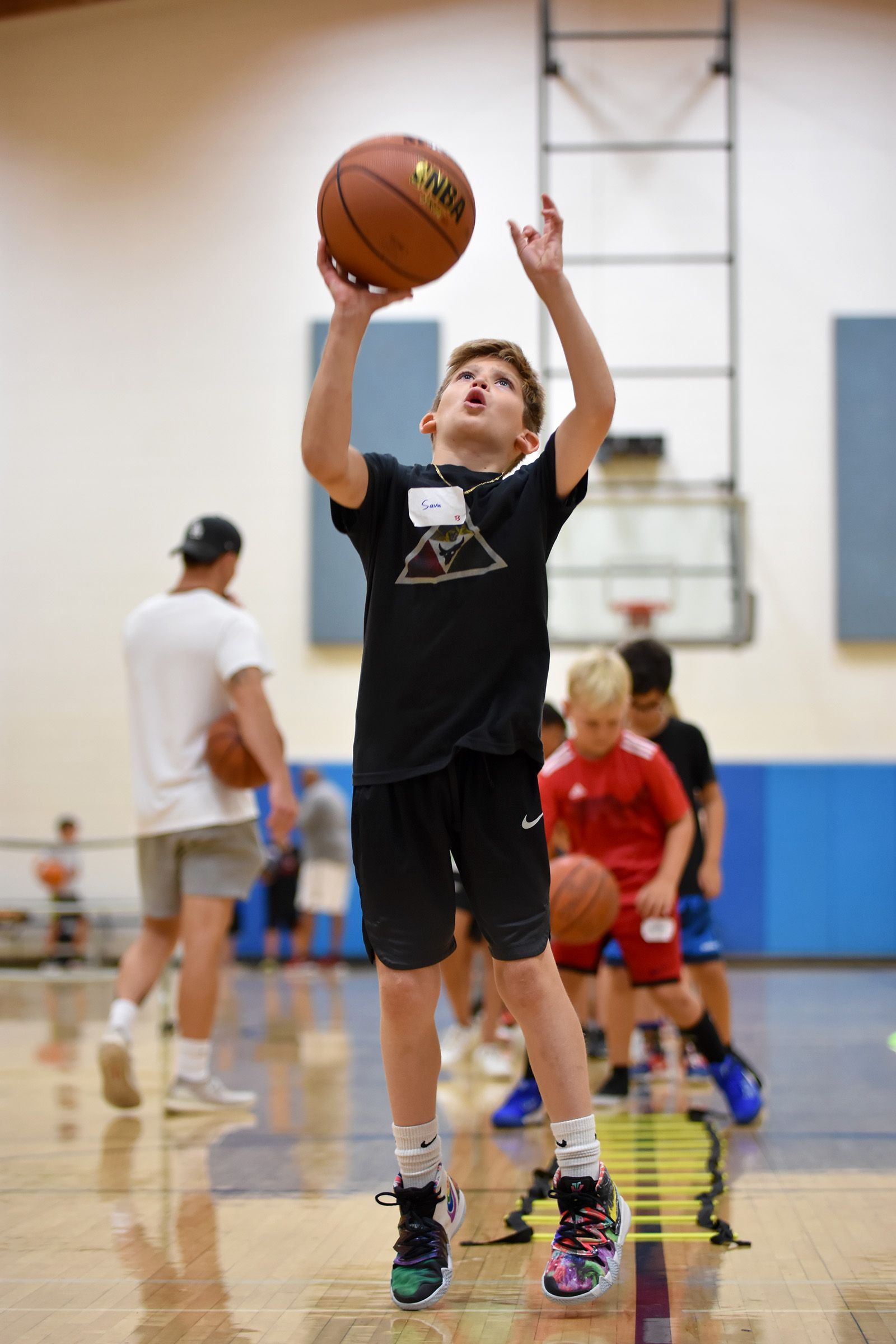 Boy reaching for a basketball during an indoor gym practice, with other players in the background