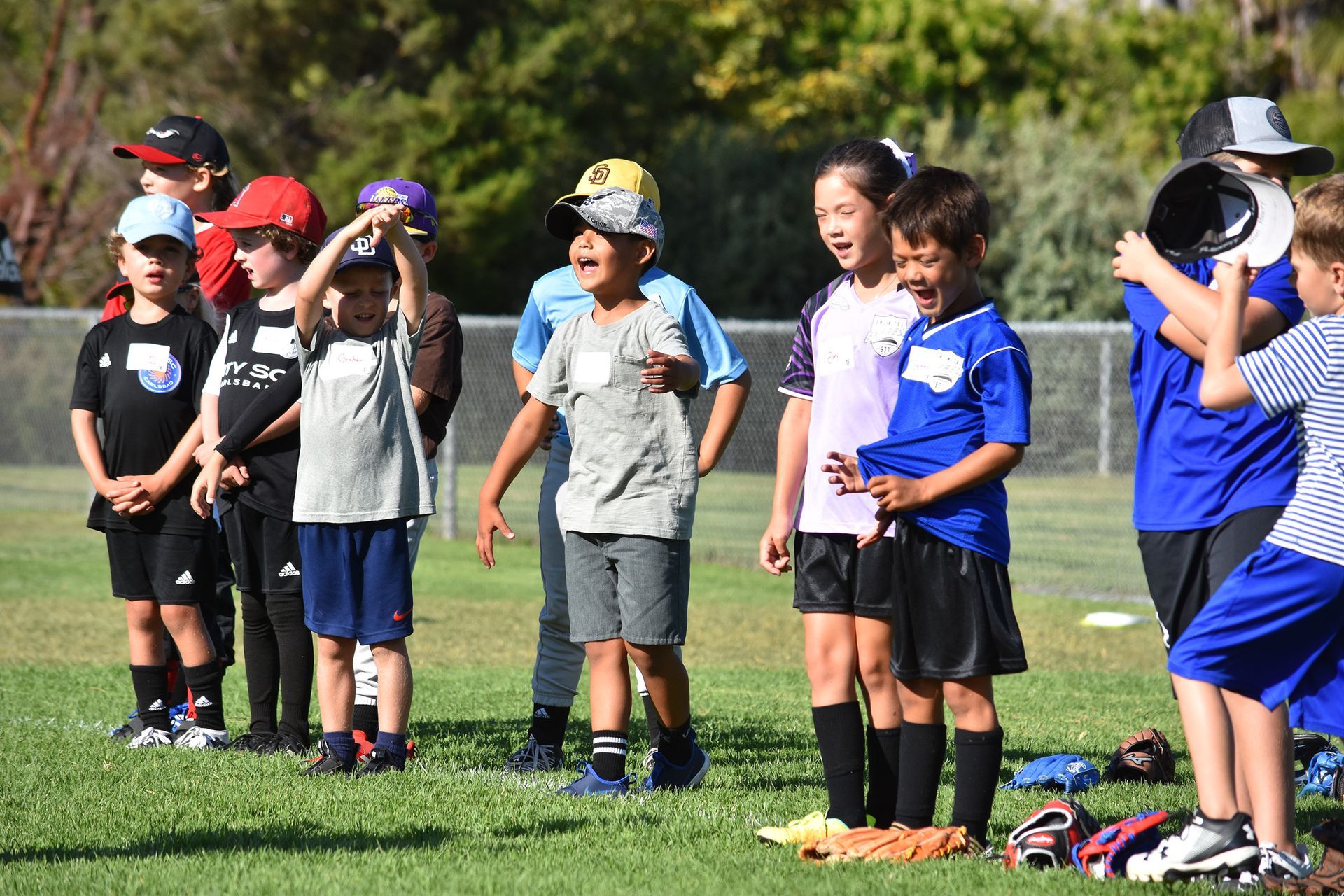 Children standing in a line on a grassy field, some in blue shirts and caps, during an outdoor event
