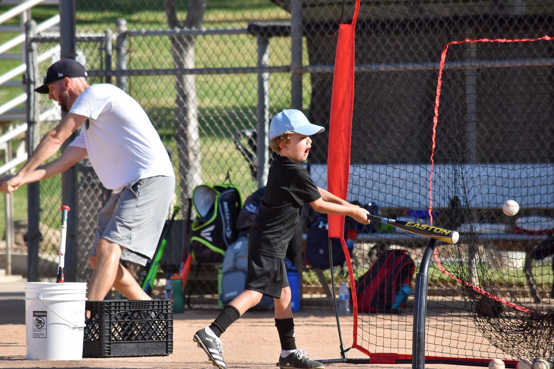 Two baseball players practice batting at a fenced field, one swinging with a coach nearby.
