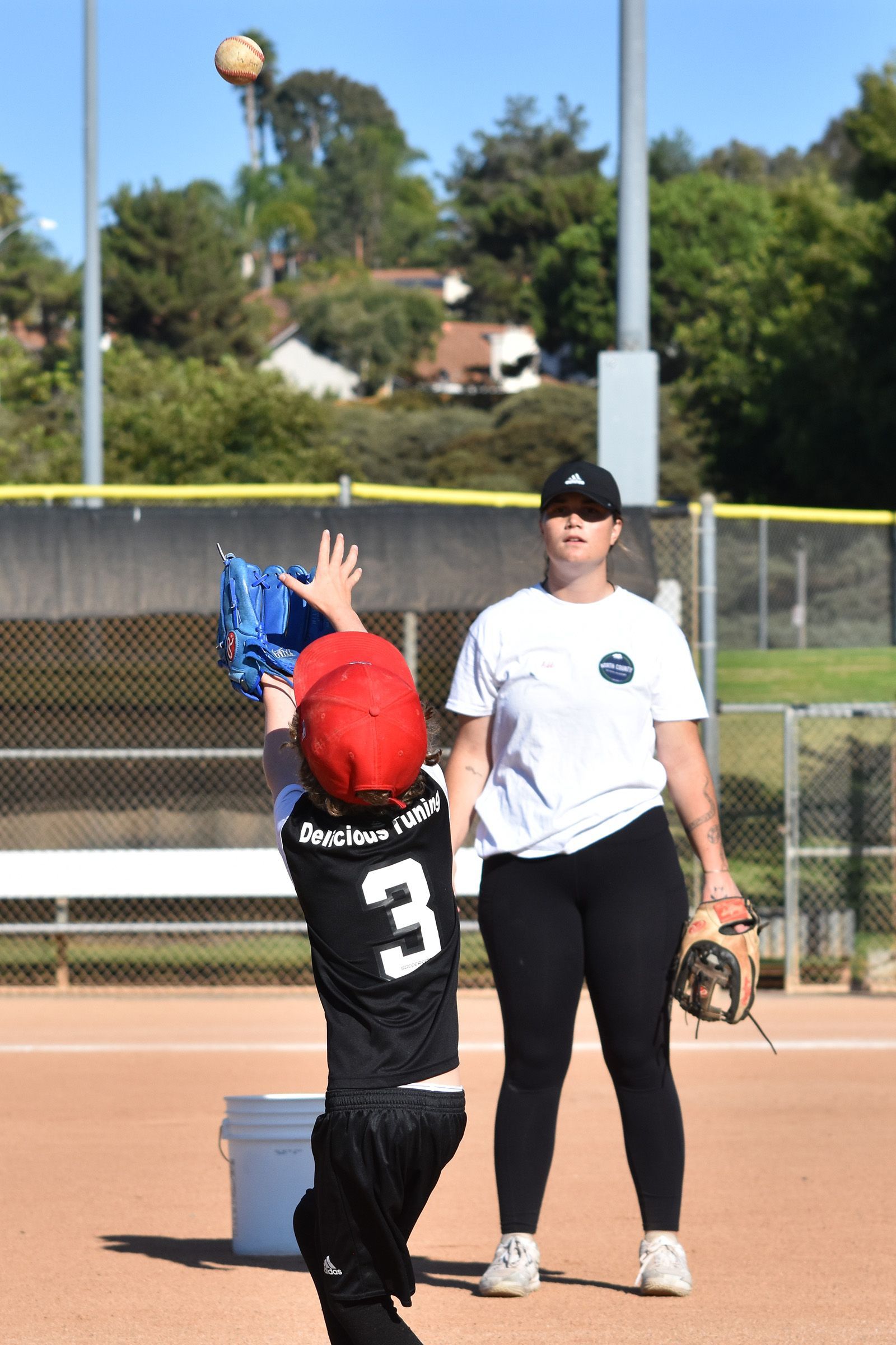Youth baseball player in black jersey reaches up for a ball near a coach on the field.