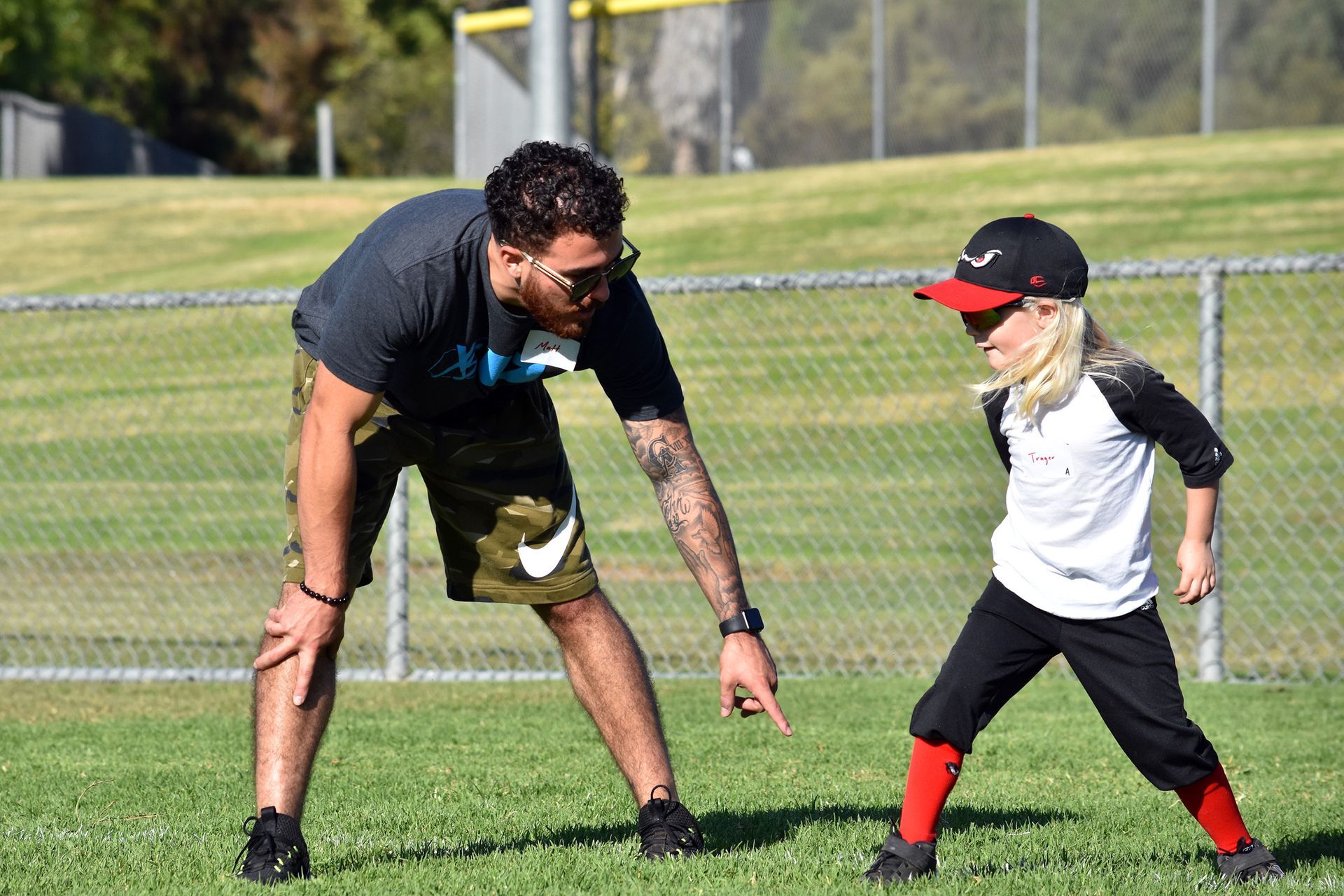 Coach and young player stretching on a grassy sports field, both leaning forward and pointing down.