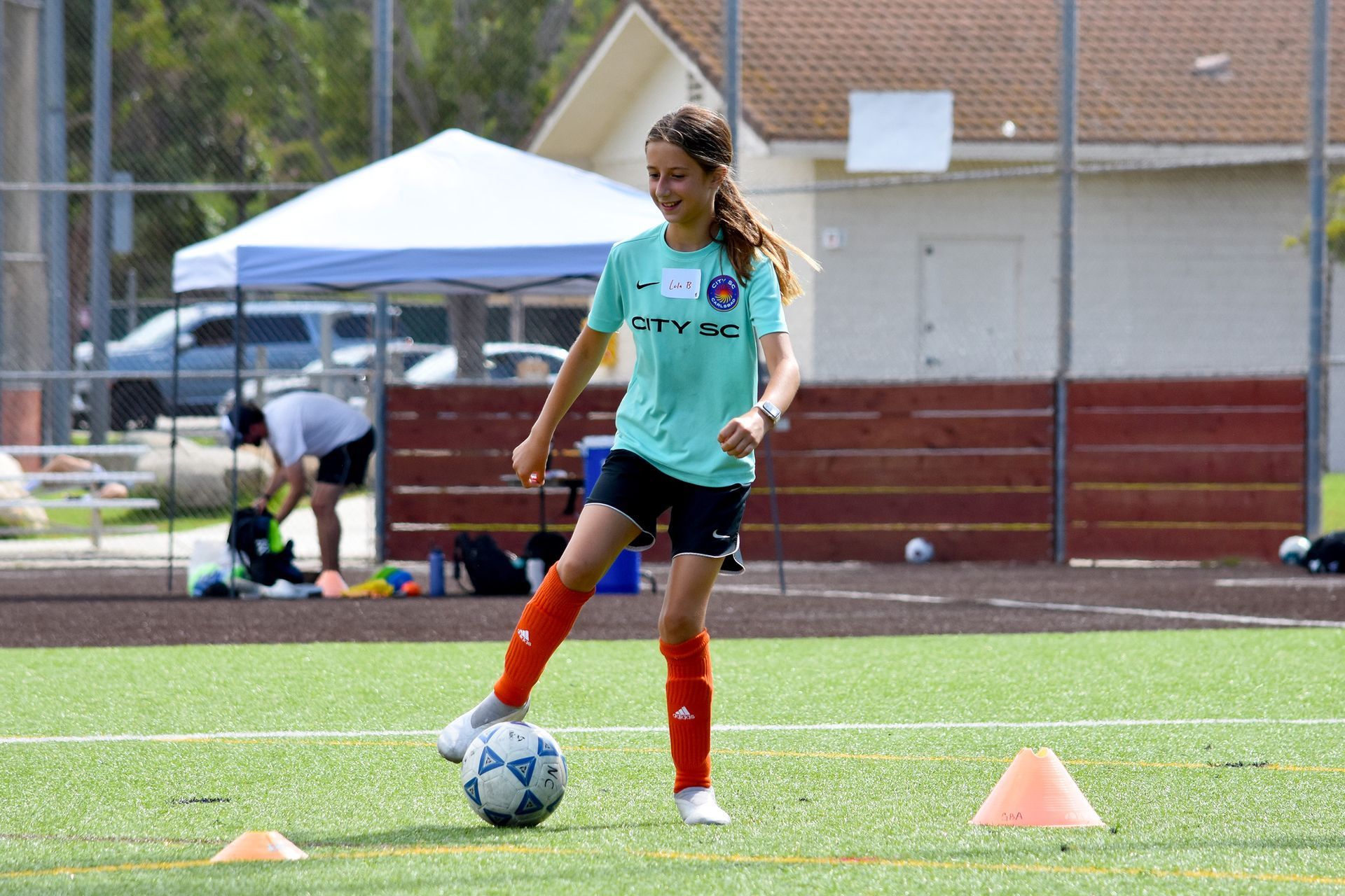Soccer player in teal shirt dribbles a ball on a field near cones and a tent.