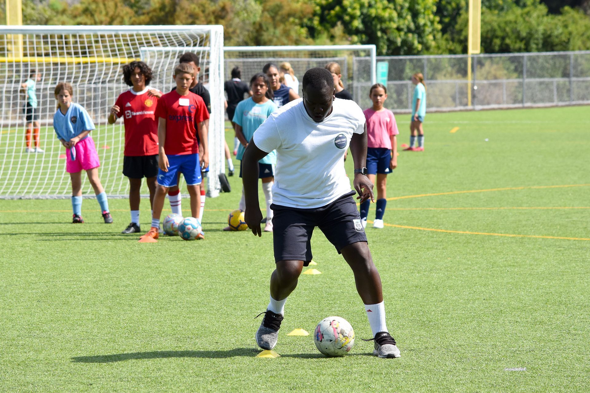 Soccer coach in white shirt instructs kids on a green field near the goal on a sunny day