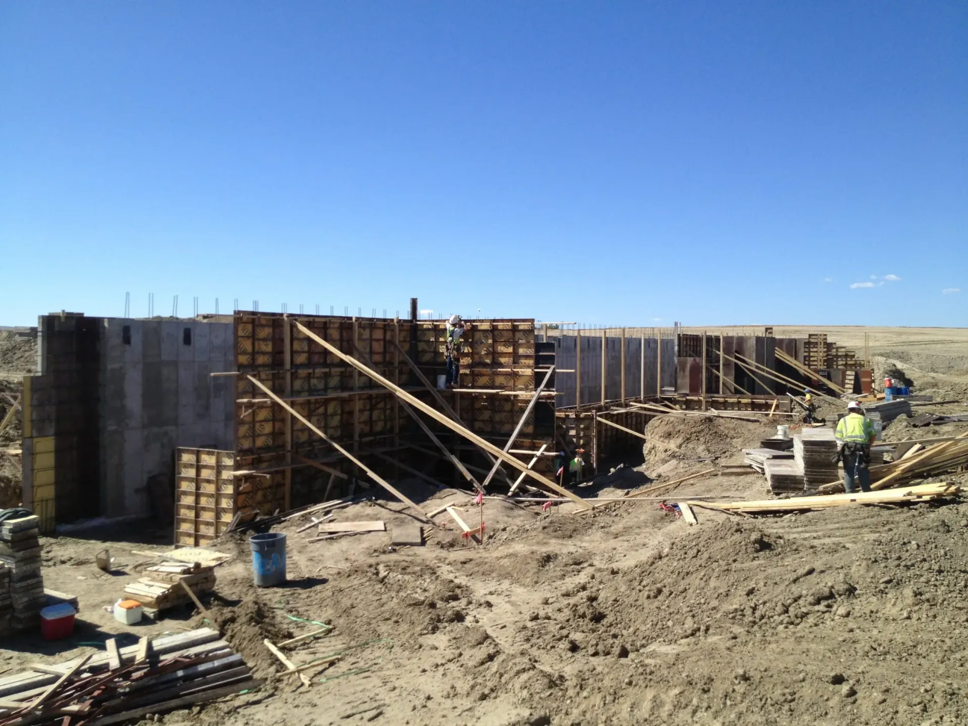 Concrete foundations with rebar in a construction site. Brown metal sheeting and red/white barriers visible.