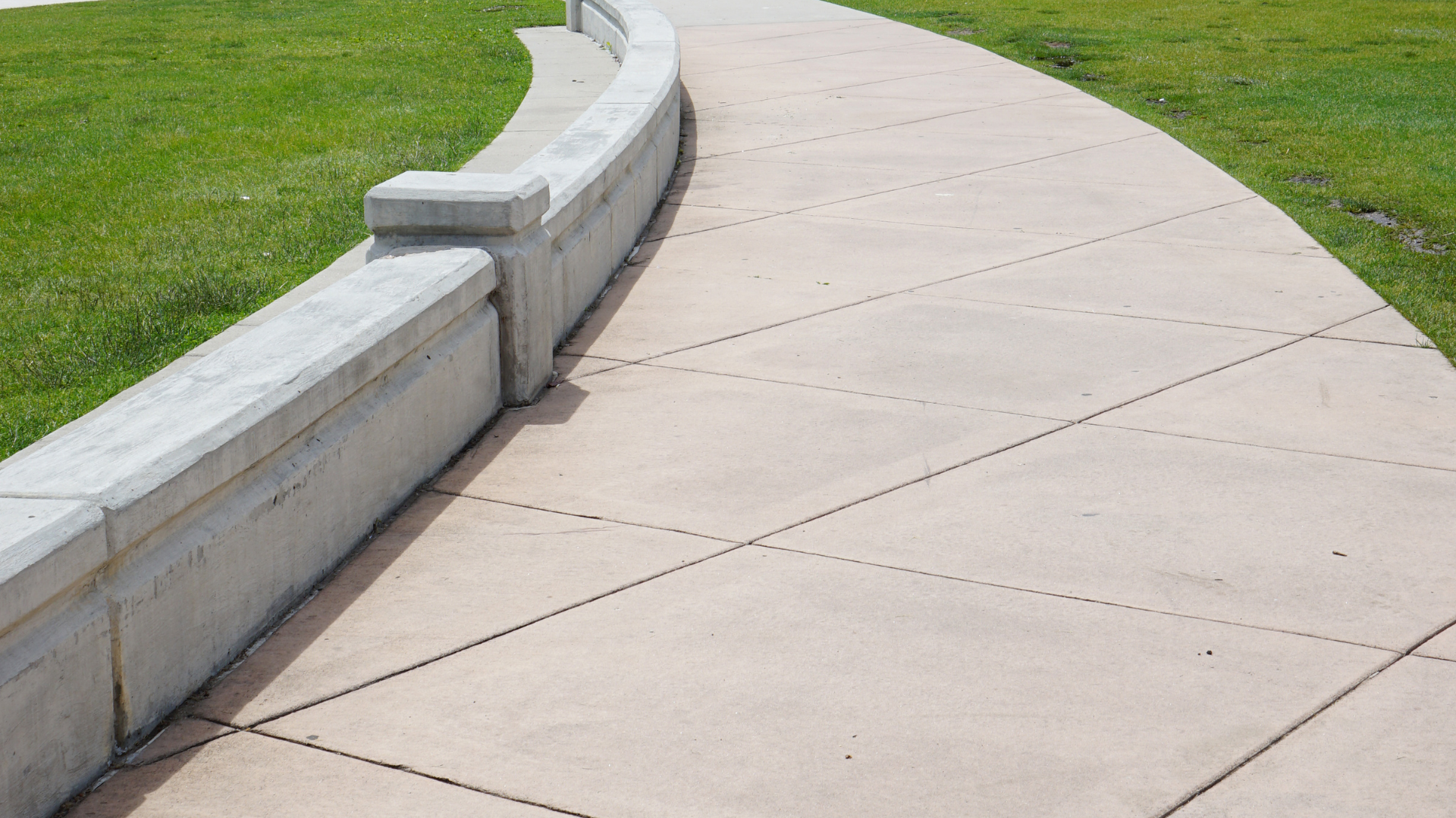 A winding sidewalk through a grassy park with houses and blue sky in the background.