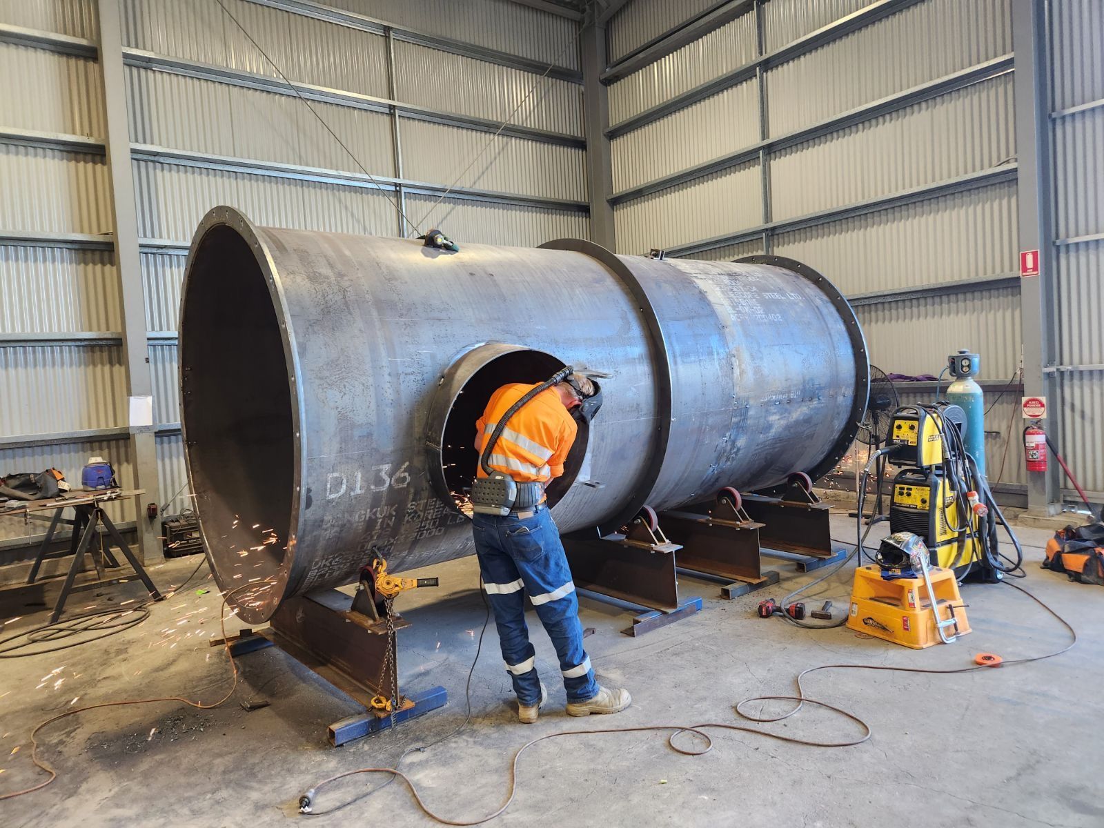 A man is welding a large metal pipe in a warehouse.