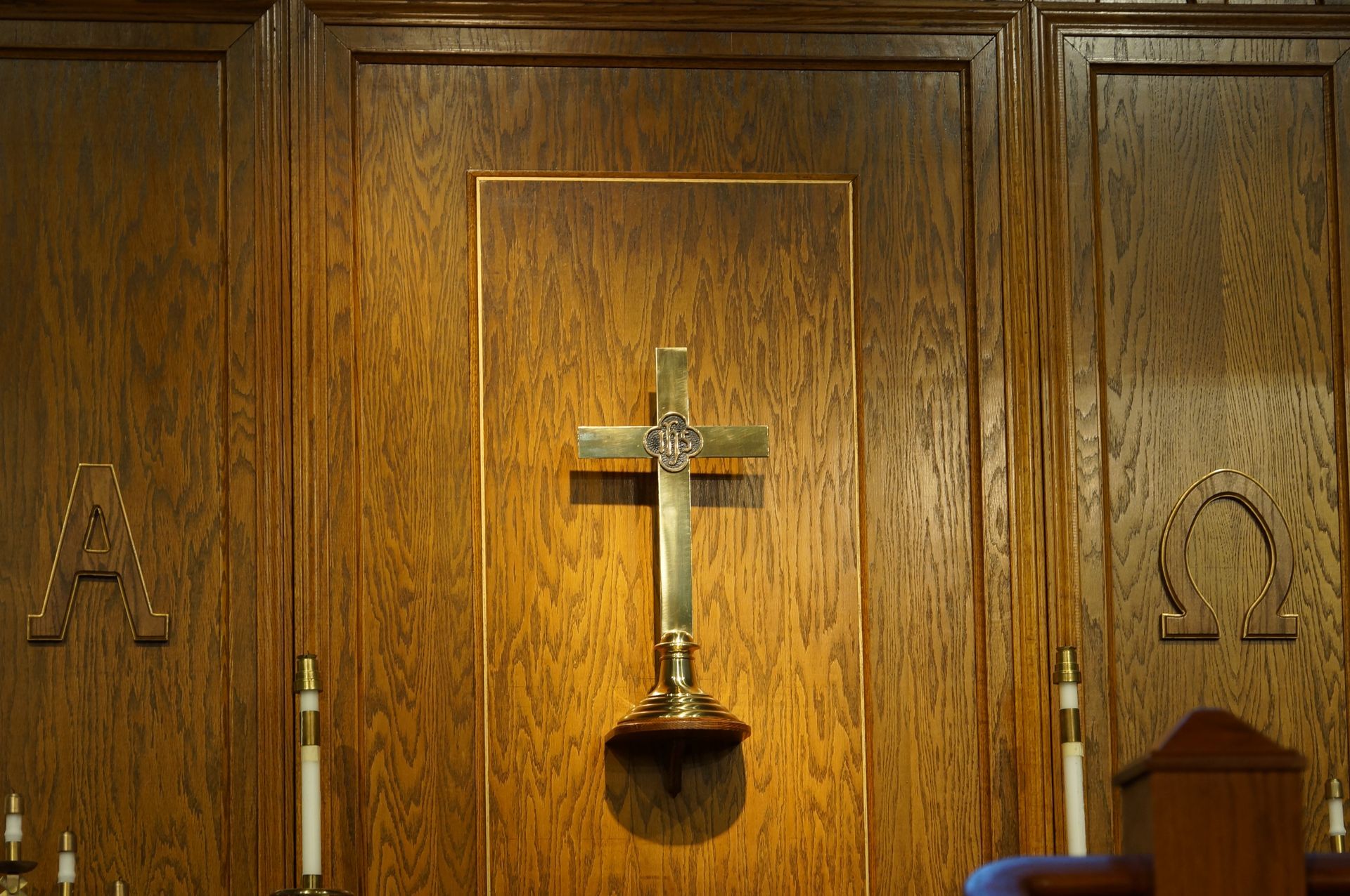 A golden cross mounted on a wooden wall between an Alpha symbol on the left and an Omega symbol on the right.