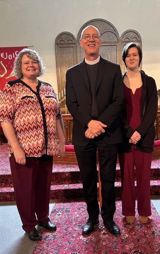 Three people stand smiling in a church before an altar, wearing formal attire, with a red banner visible on the left.