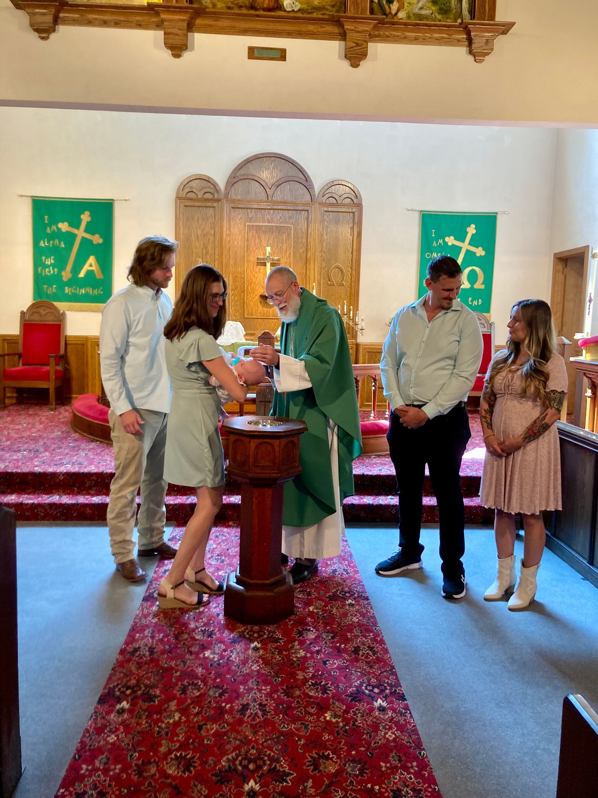 A priest performs a baptism at a font in a church, with a group of people standing around watching.