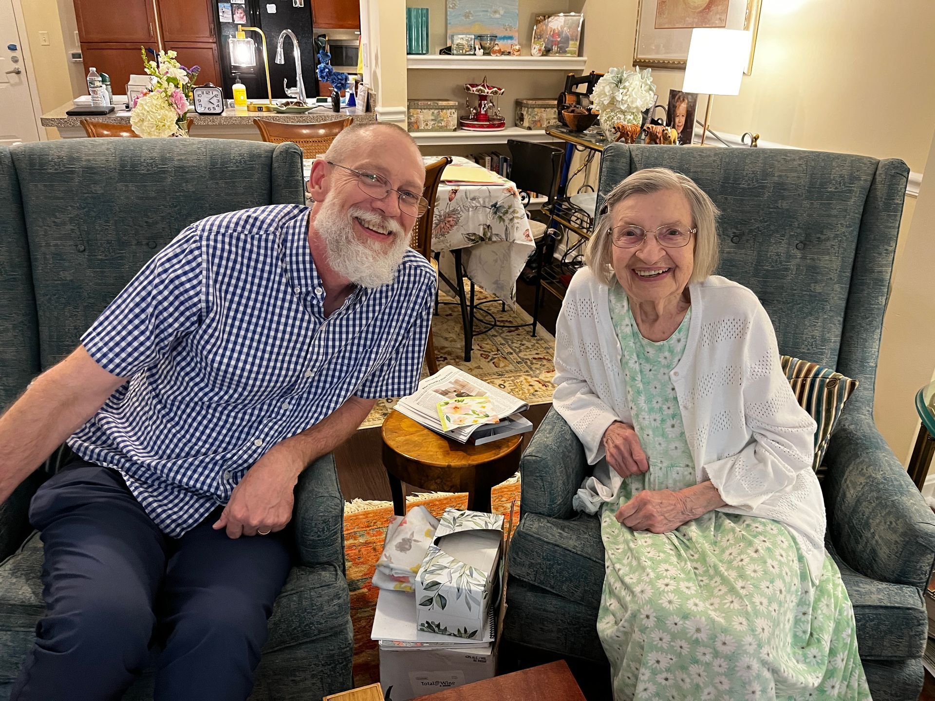 A smiling man and woman seated in upholstered armchairs in a home setting, with wrapped gifts on a table between them.