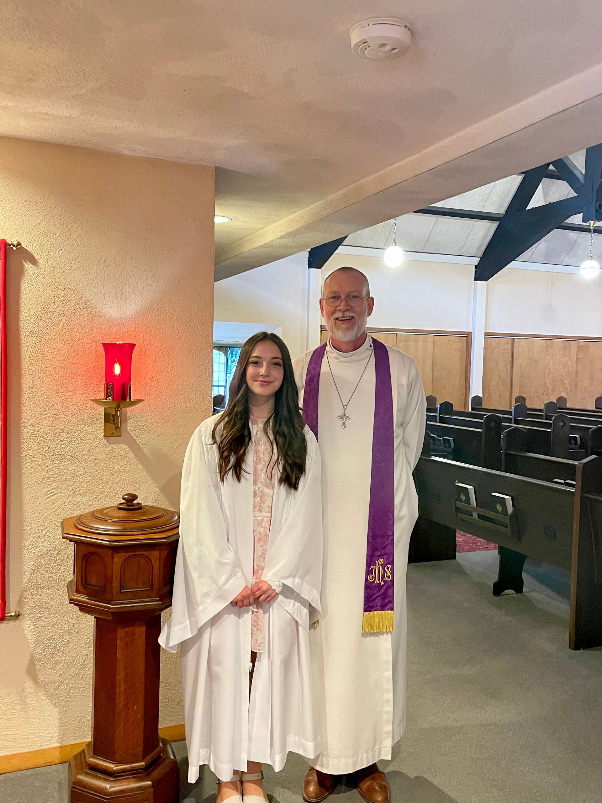 A young person and a clergyman in vestments stand together in a church next to a wooden baptismal font.