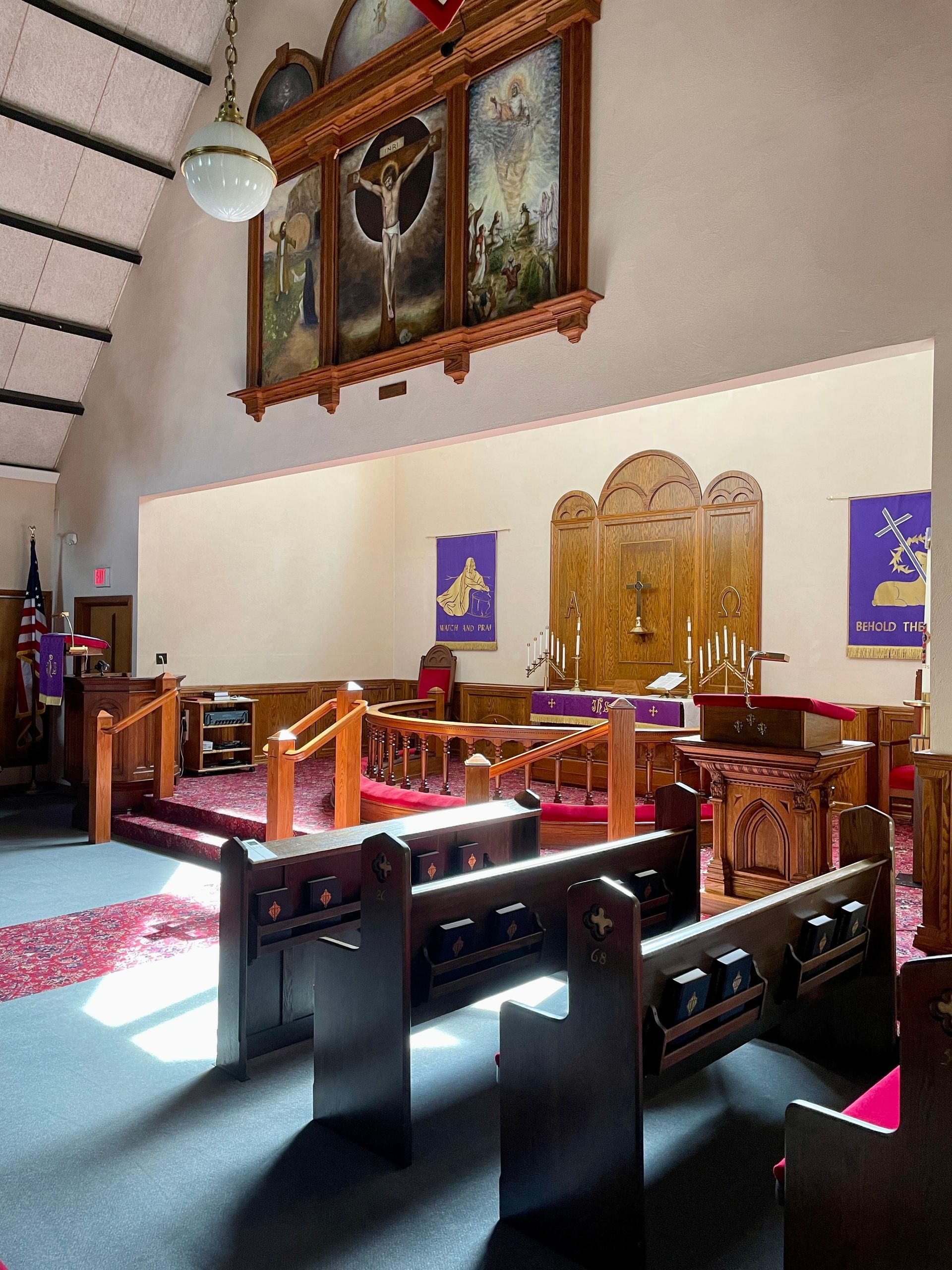 The interior of a church with wooden pews facing an altar, a decorative backdrop, and purple banners under a sloped roof.