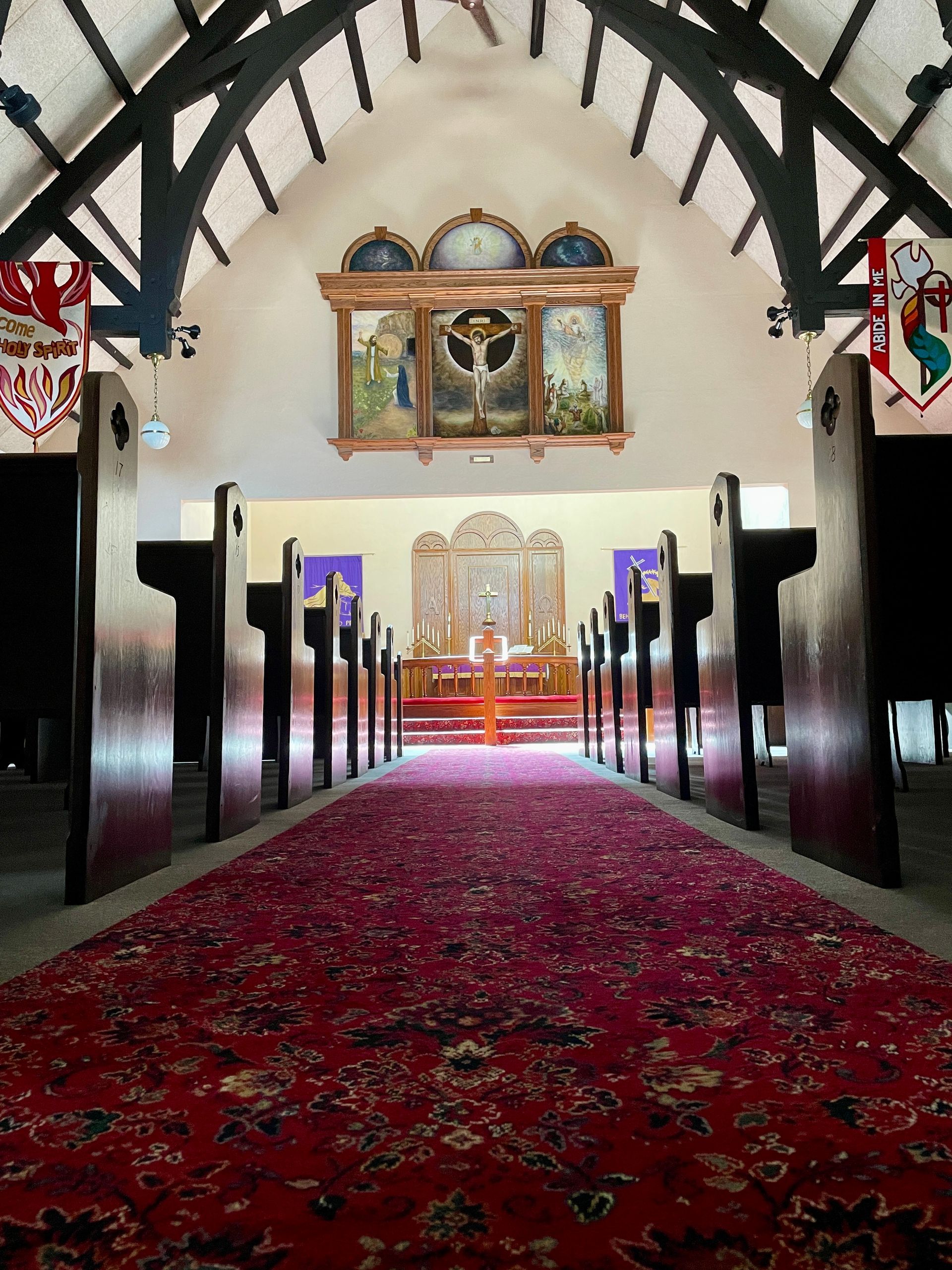 A church aisle with a long red carpet leading to an altar under a decorative wooden triptych and dark arched beams.