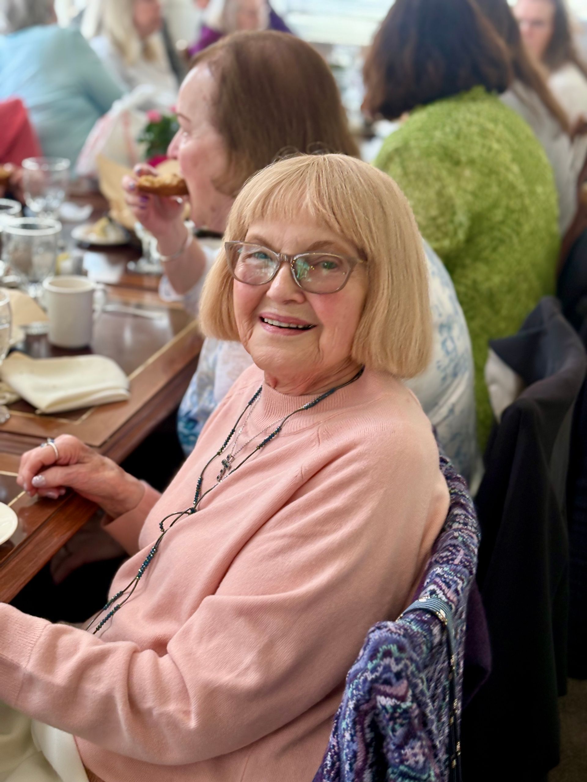 A smiling person in a pink sweater and glasses sits at a table during a group meal.