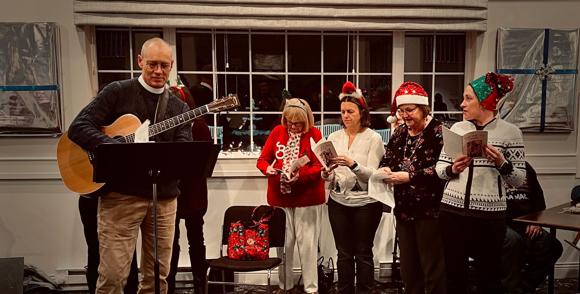 A man playing an acoustic guitar leads four people wearing holiday hats and sweaters in singing from songbooks.
