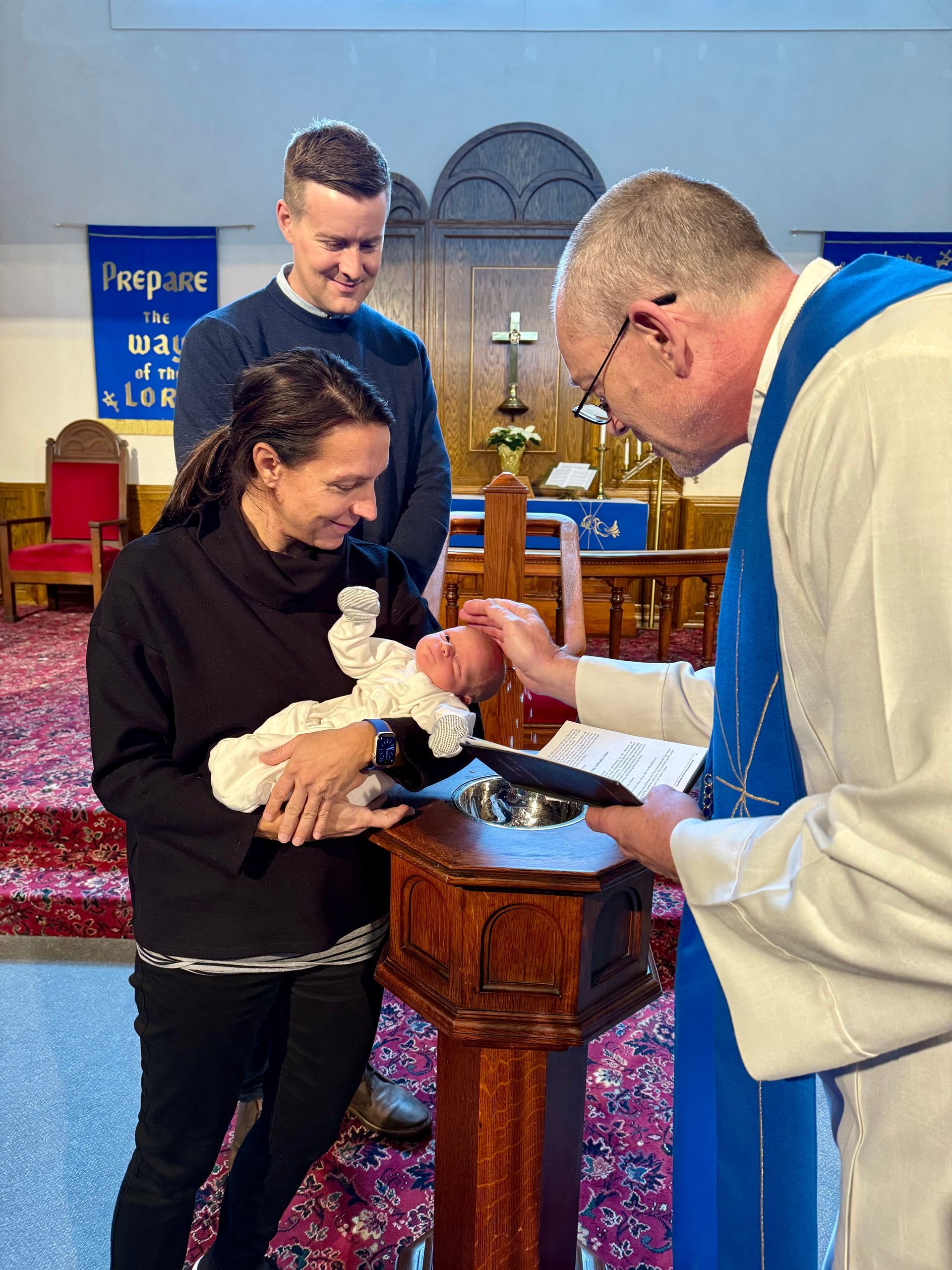 A minister performs a baptism for a baby held by a person in a church, while another person looks on with a smile.