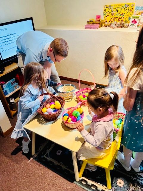 A man helping children at a table filled with baskets of colorful Easter eggs in a room with a 