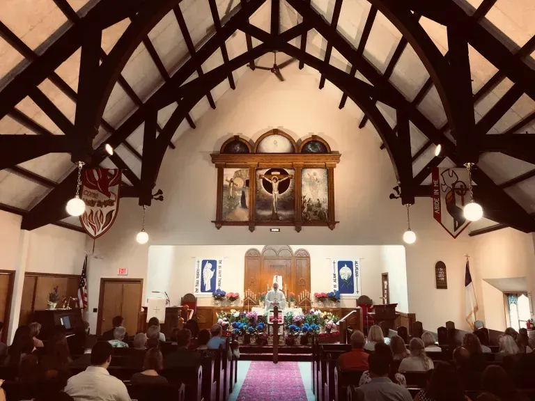 A church interior featuring a vaulted wooden ceiling, pews, a purple aisle runner, and an altar with a large triptych.