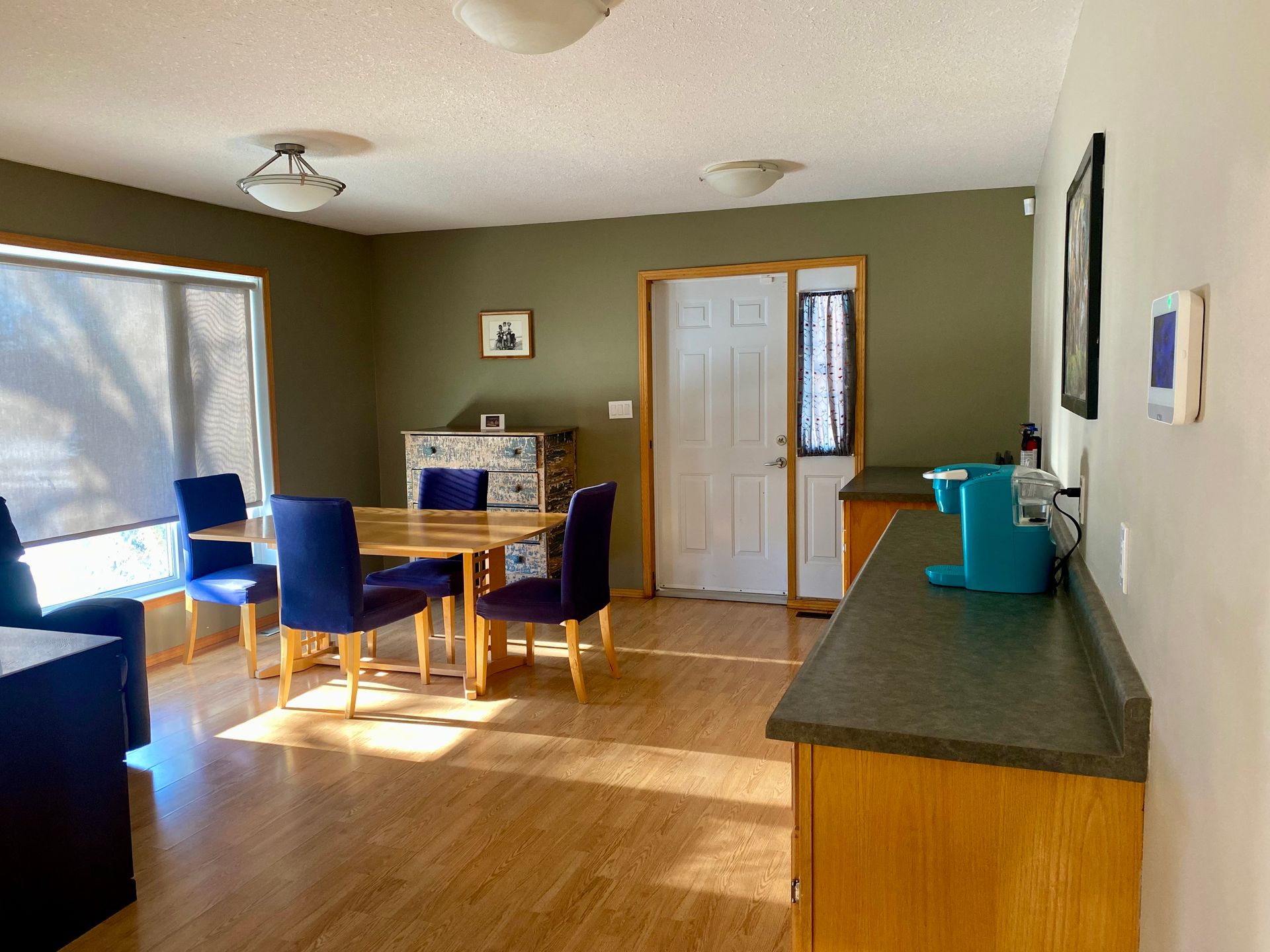 A dining area with a wood table, four blue chairs, light flooring, and a kitchen counter with a blue appliance.