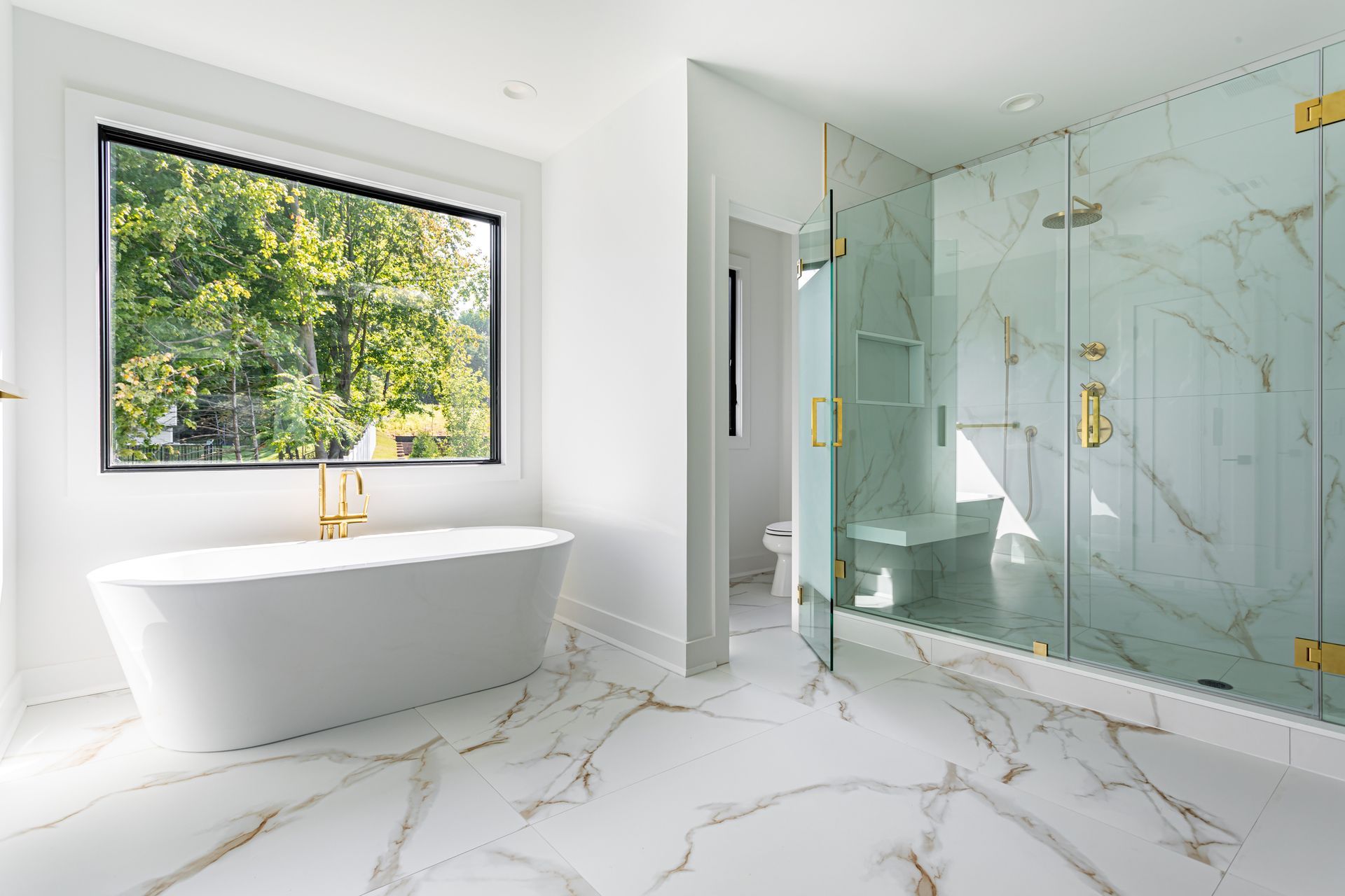 Bright modern bathroom with a freestanding tub, glass shower, and marble floors beside a large window.