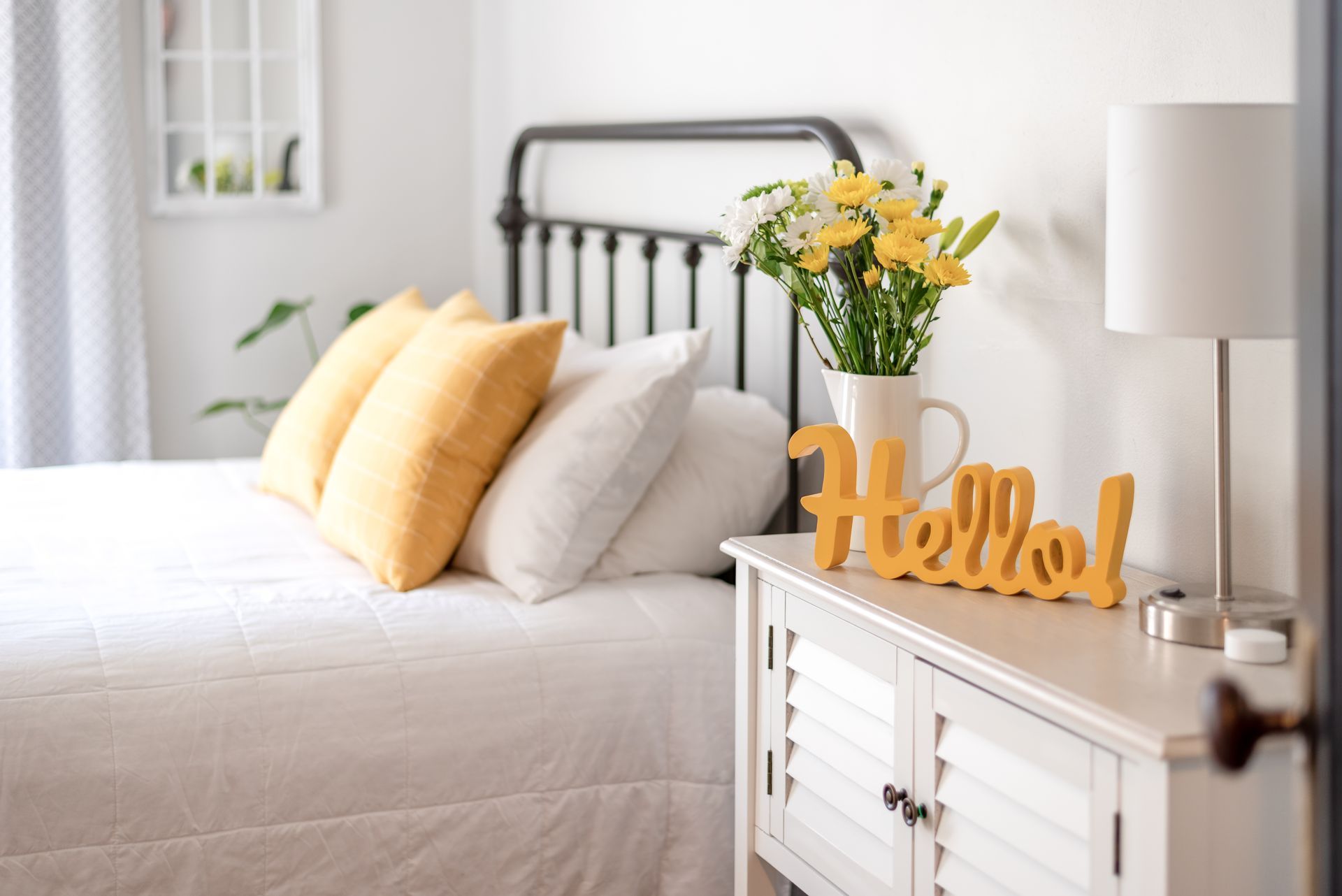 Bright bedroom with white bedding, yellow pillows, flowers, and a “Hello” sign on the dresser
