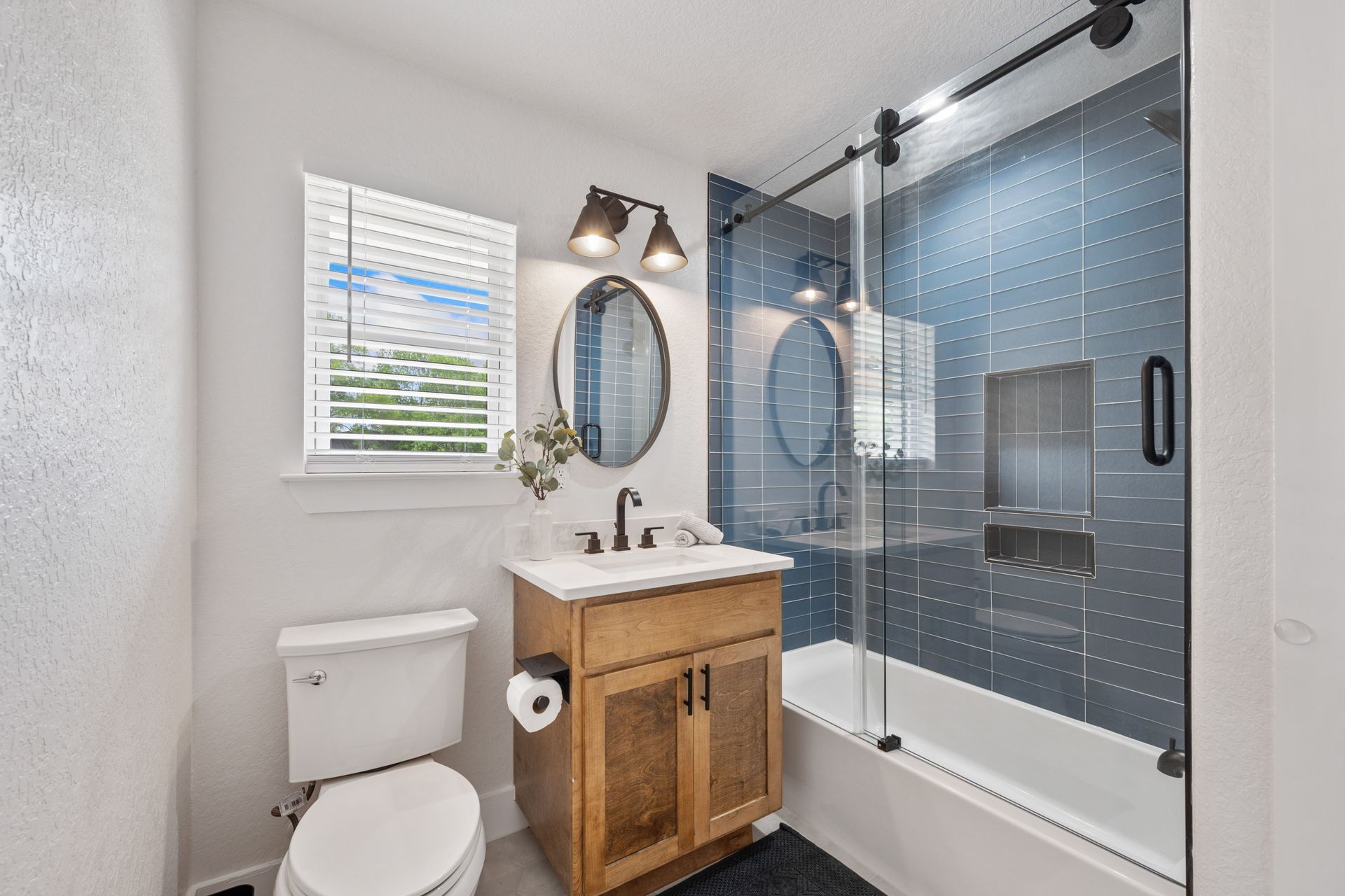 Bright bathroom with toilet, wood vanity, oval mirror, glass shower, and gray-blue tile wall.