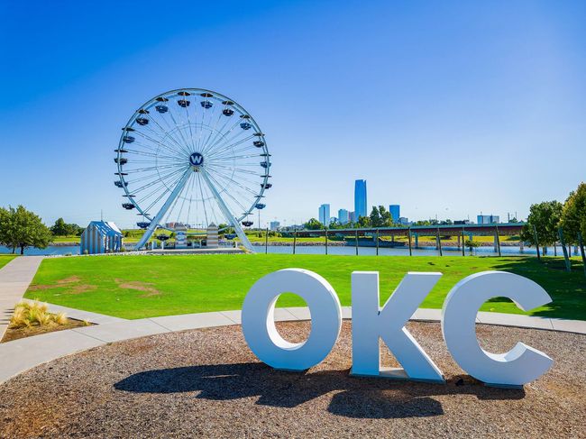 OKC sign in front of a Ferris wheel, with a city skyline in the background on a sunny day.