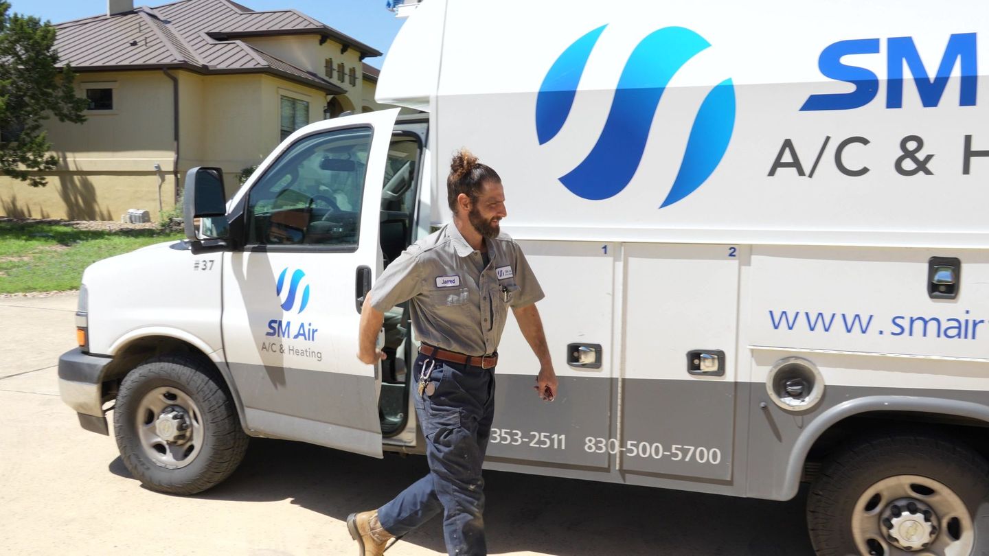 Worker walking beside a white SM A/C & Heating service truck in a sunny driveway