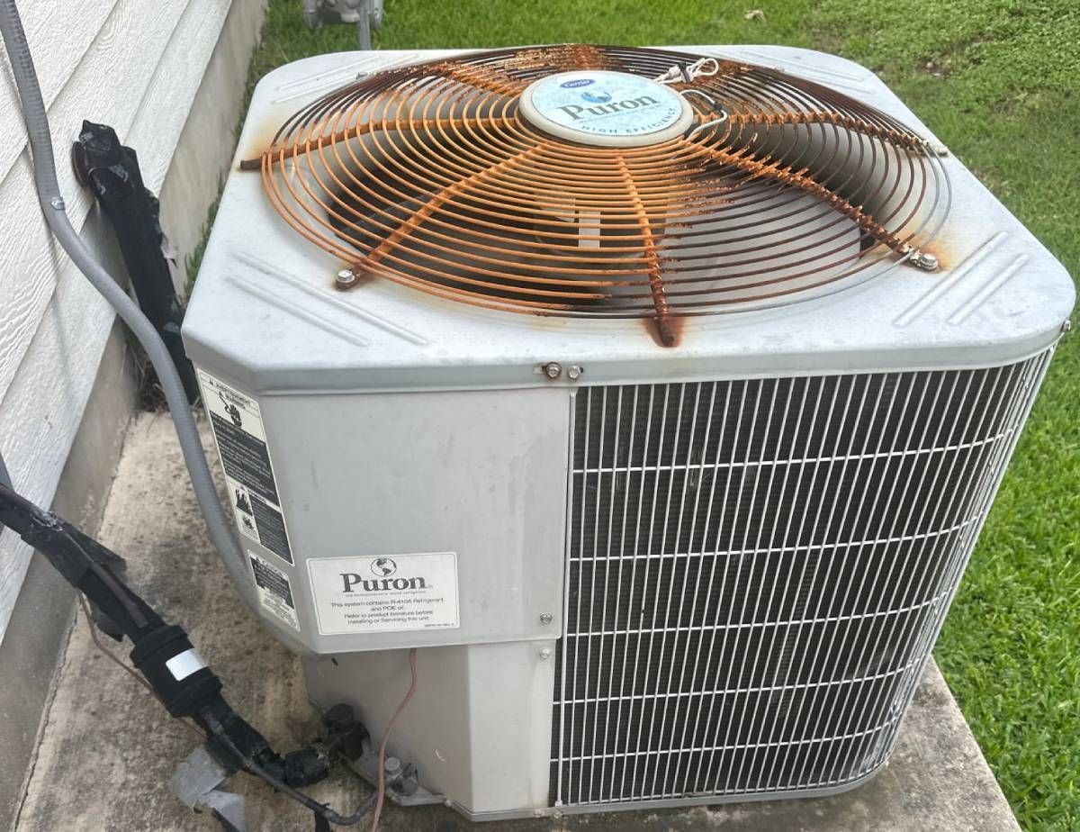 Outdoor air conditioner unit with rusted orange fan grille beside a wall on a concrete pad
