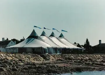 A large, white multi-peaked event tent with blue flags sits on a rocky shoreline near several houses.