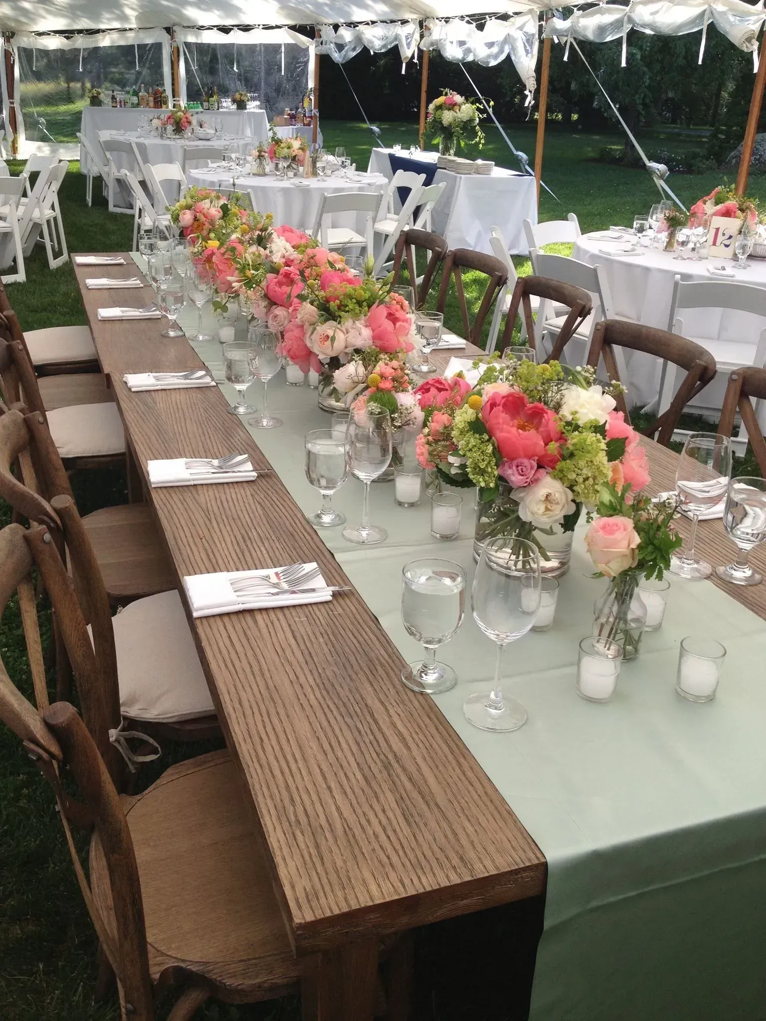 Long wooden reception table in a white event tent, decorated with pastel floral arrangements and green tablecloths.