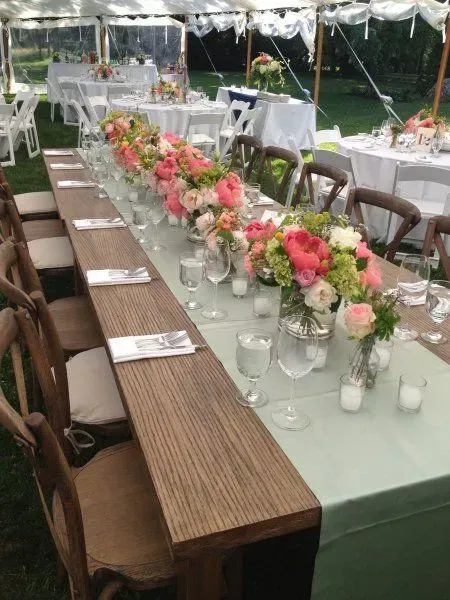 A long wooden banquet table under a white tent, decorated with floral centerpieces, greenery, and glassware for a party.