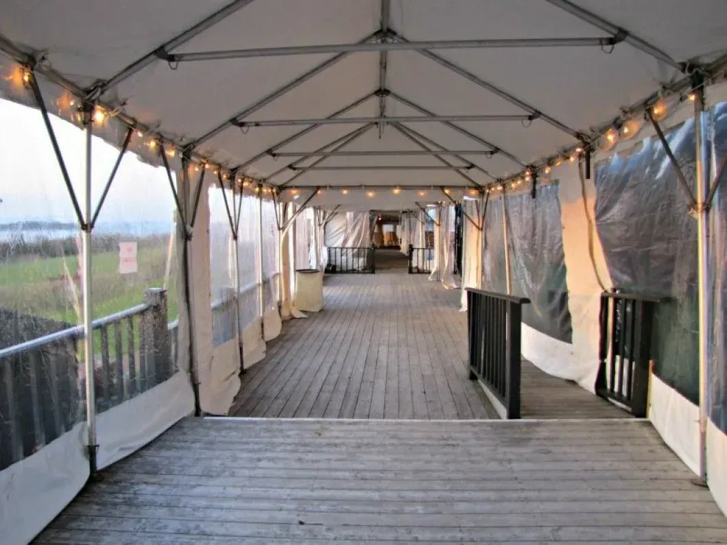 A covered wooden walkway lined with lights, enclosed by clear plastic walls, leading to a distant outdoor area.