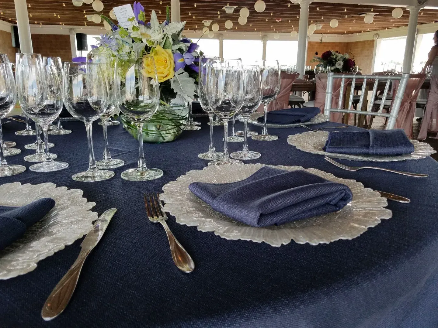 A navy blue tablecloth is set with silver chargers, dark blue napkins, cutlery, and wine glasses, with floral centerpieces.