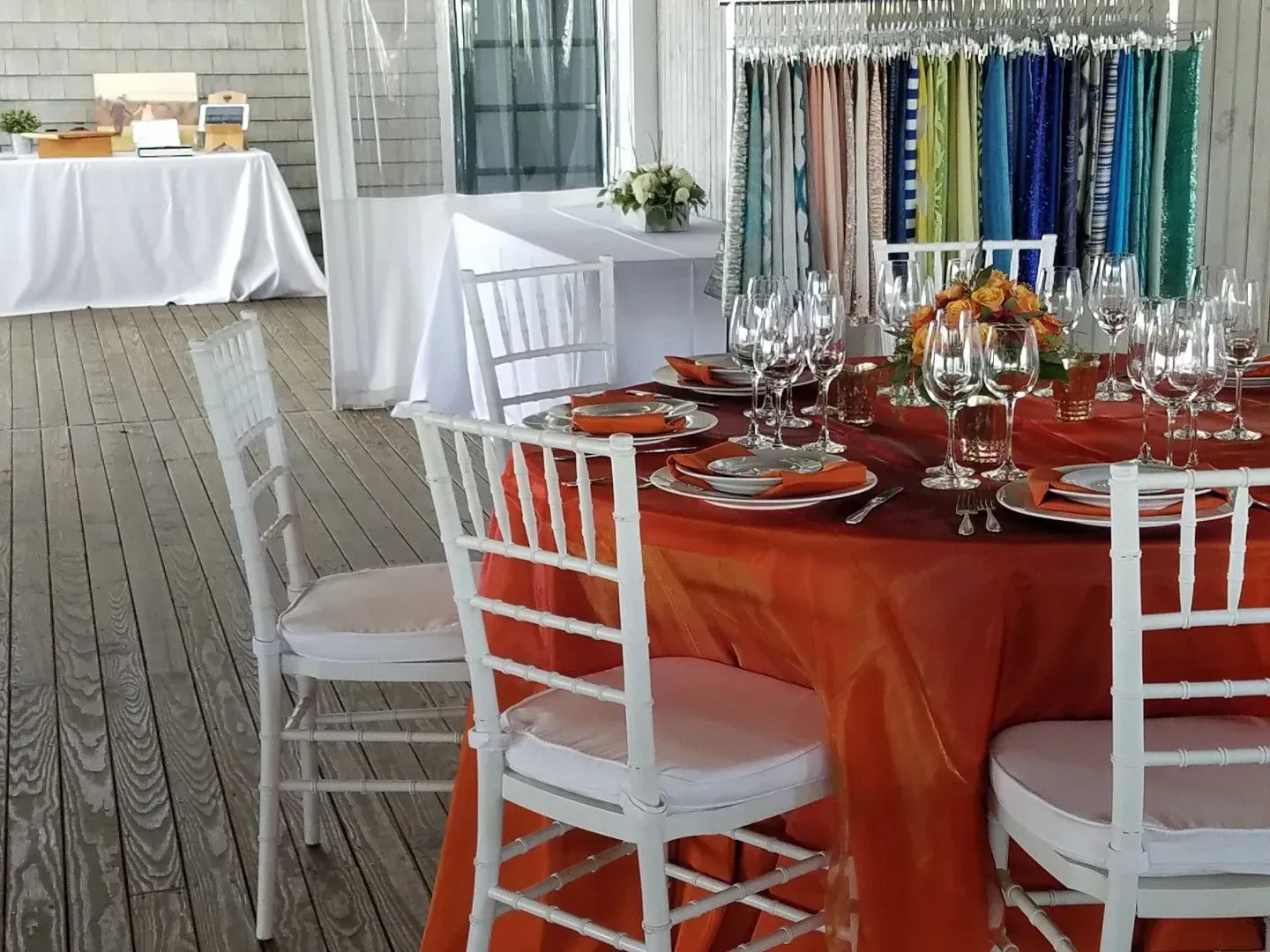 A round table with an orange tablecloth and white chairs set for an event, with fabric samples hanging in the background.