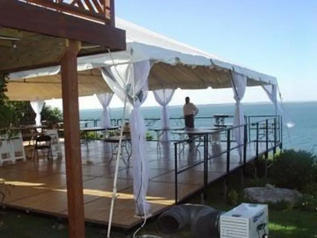 A white tent on a wooden deck overlooks the ocean, featuring sheer curtains and a person standing near the railing.