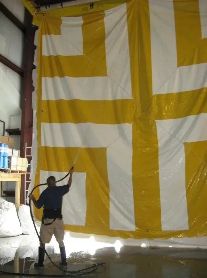 A worker in boots uses a high-pressure hose to clean a large yellow and white vinyl curtain in an industrial warehouse.