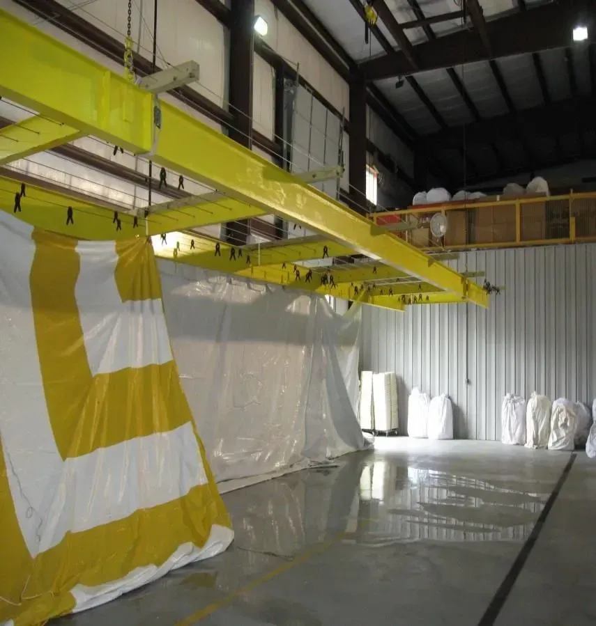 A large warehouse interior featuring yellow overhead beams supporting white and yellow plastic curtains hanging on tracks.