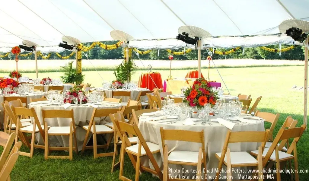 Round tables with white tablecloths and wooden chairs set up for a wedding or event inside a tent on a grassy field.