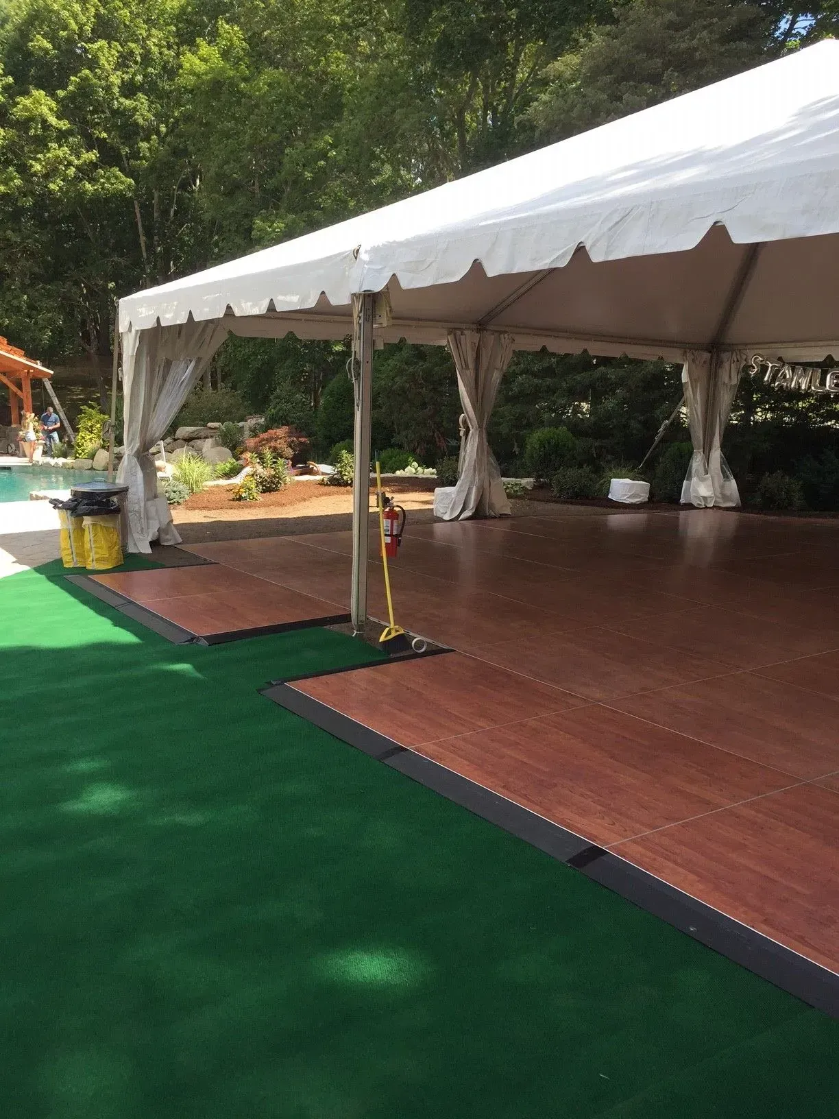 A large white party tent covers a polished wood dance floor, set on a bright green carpeted area outdoors.