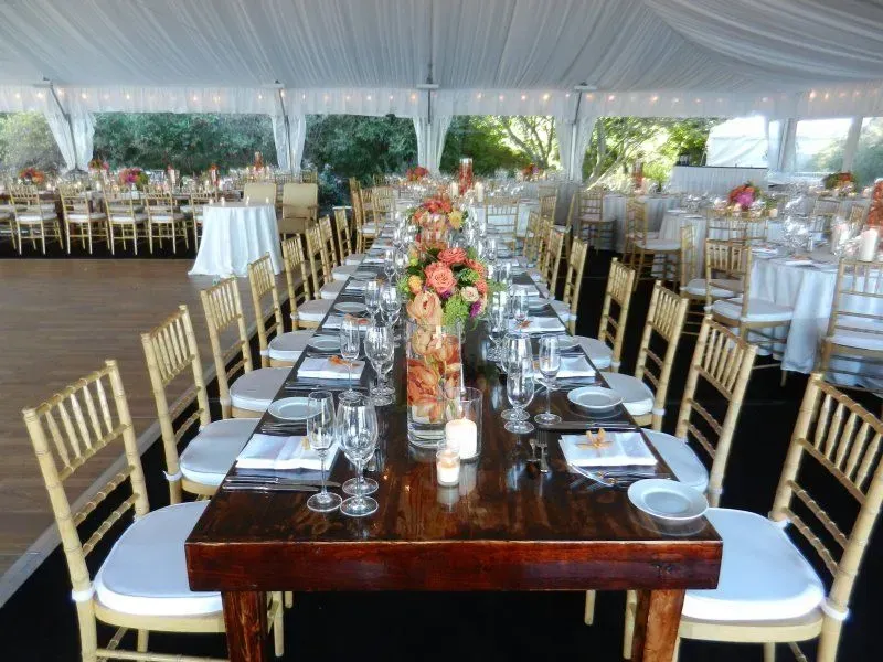 Long wooden dining table with floral centerpieces and bamboo-style chairs arranged for an outdoor tent wedding reception.