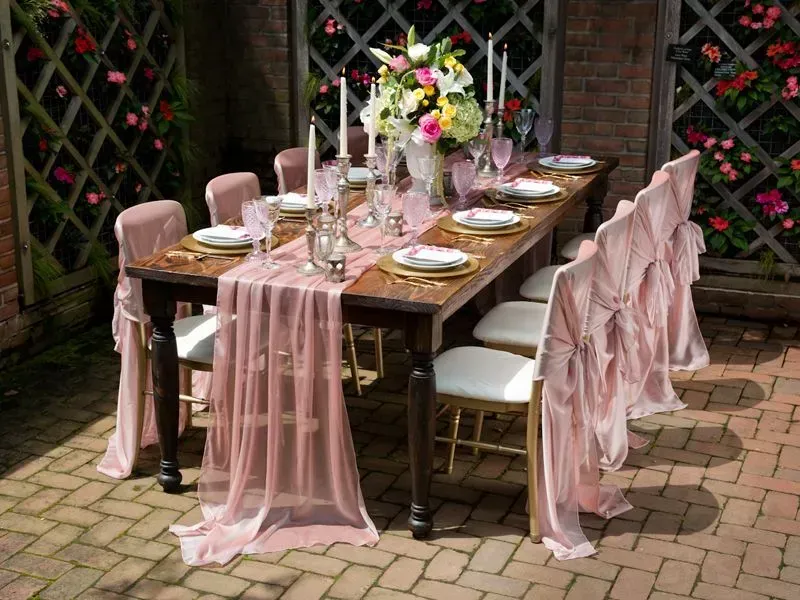 A formal dining table set for an outdoor event, featuring a dusty rose chiffon runner, matching chair covers, and flowers.
