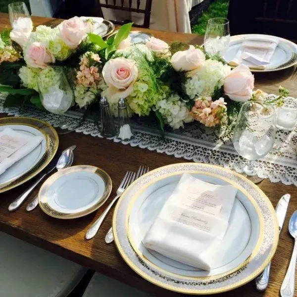 An elegant wedding table setting with gold-rimmed plates, lace runners, and a centerpiece of light pink roses and hydrangeas.