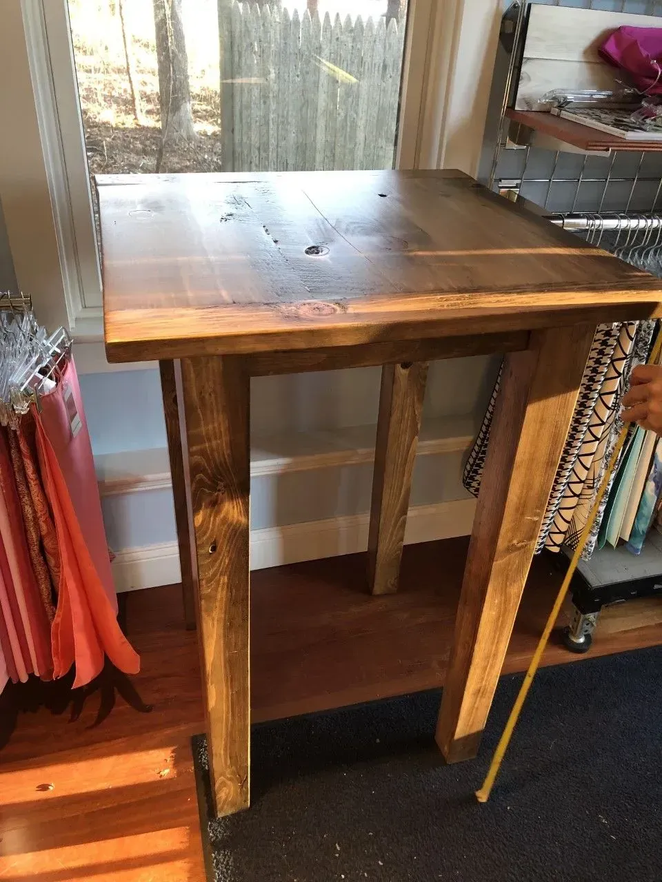 A rustic, dark-stained wooden high-top table stands indoors near a window, next to hanging fabric samples.