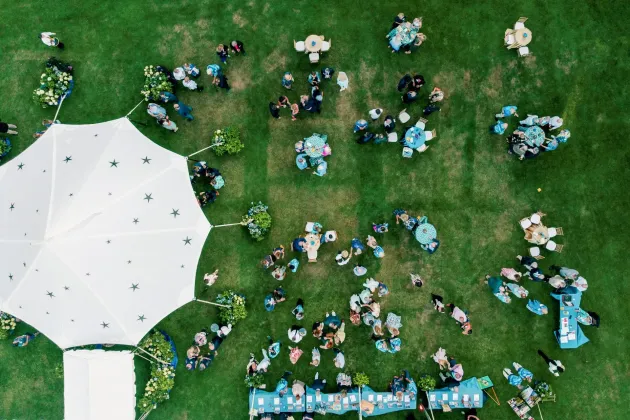 An aerial view of an outdoor event with a large white tent, guests mingling, and tables set up on a green lawn.