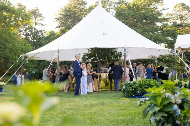 People socialize under a large white event tent on a grassy lawn with trees and hydrangea bushes in the background.