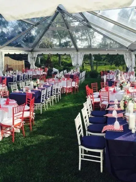 Outdoor wedding reception tent with rows of tables set with navy tablecloths, red accents, and white chairs on grass.
