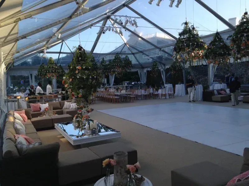A large tent with a glass roof and white dance floor, set up for an event with lounge seating and hanging floral decor.